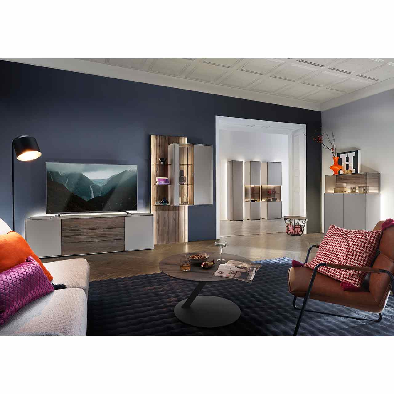 Wohnzimmer mit Wohnwand bestehend aus einem Lowboard in dunkelbraunem Holz und Lack sand, einer hellen Hängevitrine eingefasst in ein beleuchtetes Wandpaneel aus Altholzbuche mit Glasböden. Davor runder Couchtisch, rechts passendes Highboard.
