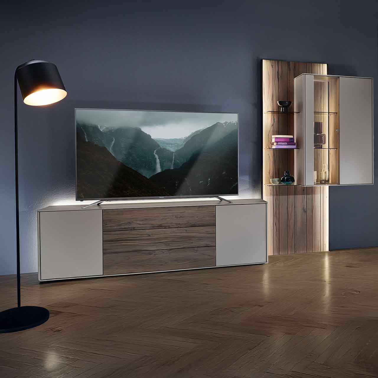 Hartmann AUDRA 5610 - Wohnwand Nr. V26 - dreiteilig aus Altholzbuche Uyaa und Lack Sand Modernes Wohnzimmer mit dreiteiliger Wohnwand bestehend aus einem Lowboard in dunkelbraunem Holz und Lack sand, einer Hängevitrine in Lack sand eingefasst in ein beleuchtetes Wandpaneel aus Altholzbuche mit Glasböden.