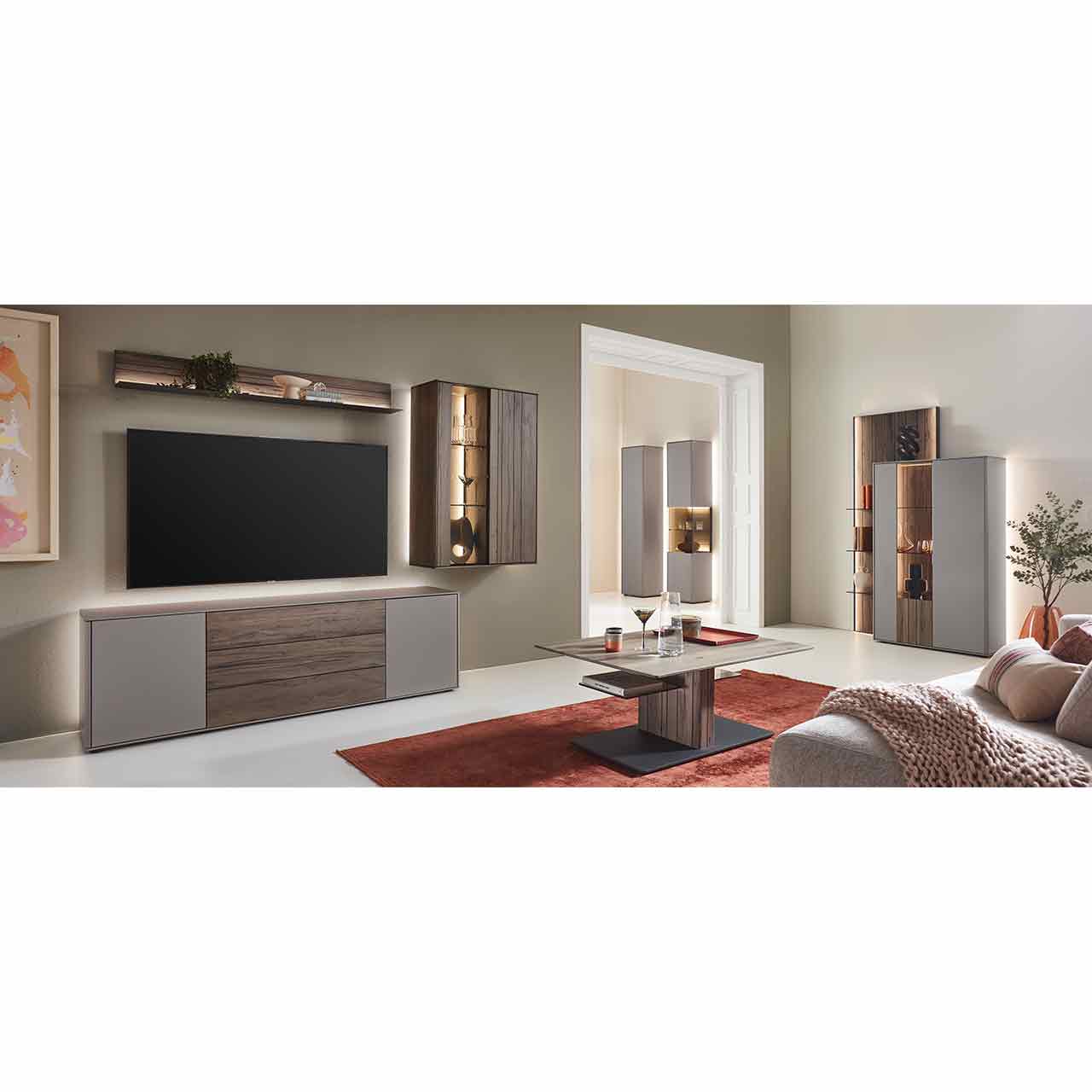 Wohnzimmer mit moderner Massivholzwohnwand bestehend aus einem Lowboard in dunkelbraunem Holz und Lack sand, einer beleuchteten Hängevitrine und einem Wandregal in dunkelbraun. Davor rechteckiger Couchtisch, rechts passendes Highboard an Wandpaneel.