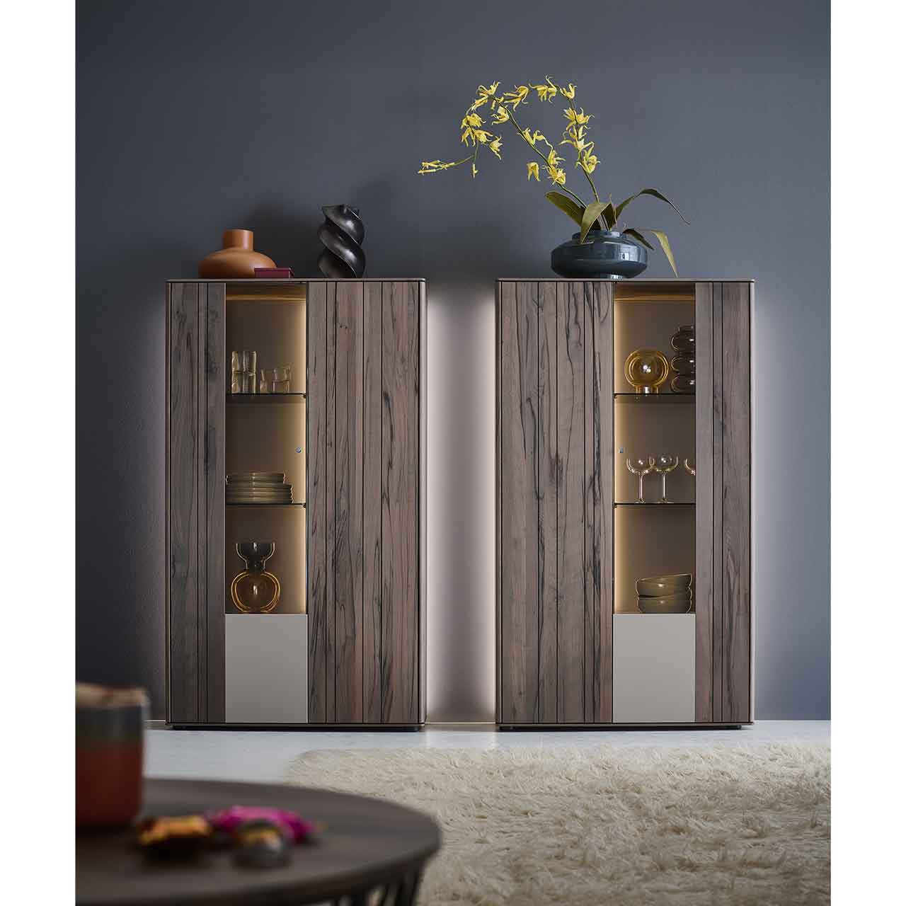 Wohnzimmer mit zwei modernen Highboards aus massiver, dunkelbrauner Altholzbuche Uyaa, jeweils mit beleuchtetem Glasausschnitt und Glasböden, vor beleuchteter Rückwand in lack Sand.