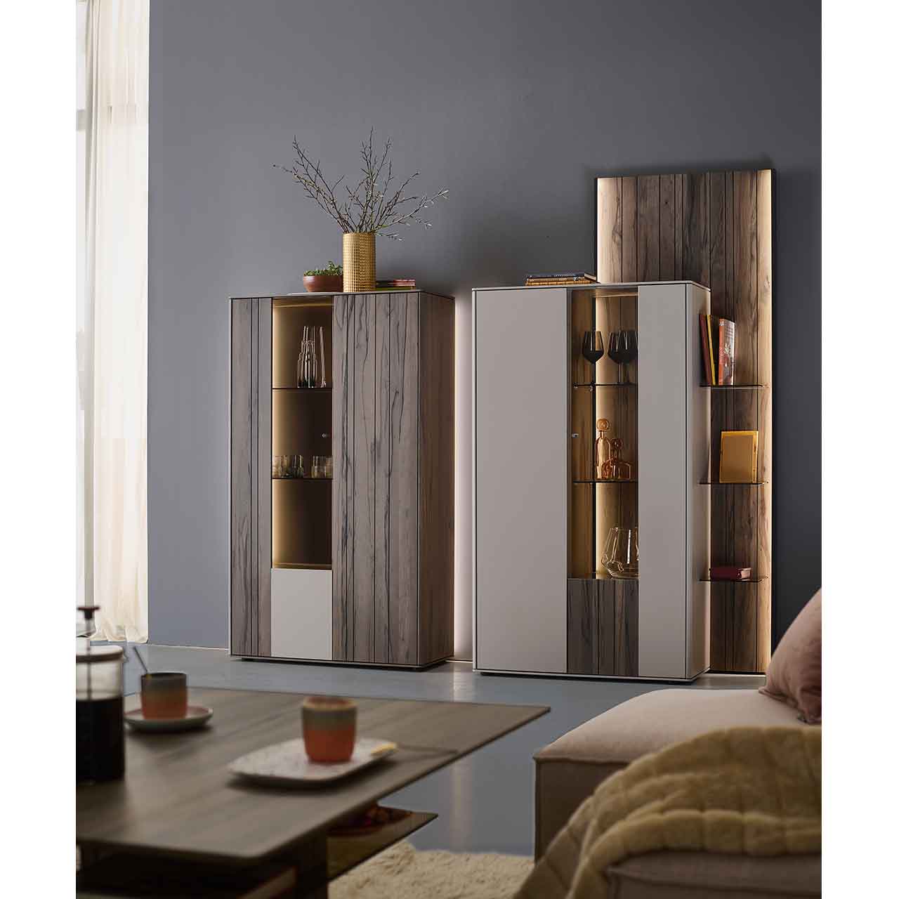 Wohnzimmer mit zwei modernen Massivholz-Highboards in sandfarbenen Lack und dunkelbrauner Altholzbuche Uyaa, jeweils mit beleuchtetem Glasausschnitt und Glasböden, ergänzt durch ein seitliches, beleuchtetes Holzwandpaneel.
