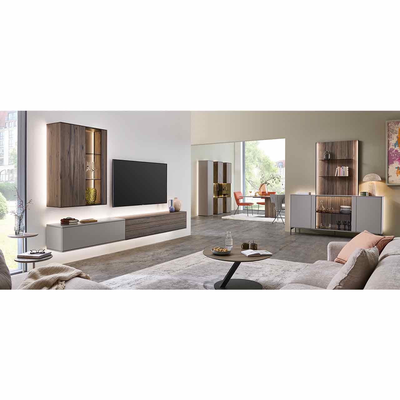 Wohnzimmer mit moderner Massivholzwohnwand bestehend aus zwei Lowboards in dunkelbraunem Holz und Lack sand und beleuchteter Hängevitrine in dunkelbraun. Davor runder Couchtisch, rechts passendes Sideboard an edlem Wandpaneel mit Glasböden.