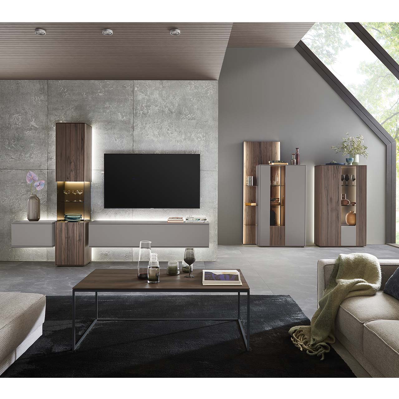 Modernes Wohnzimmer mit dreiteiliger Wohnwand bestehend aus einer Standvitrine in dunkelbraunem Holz mit Maserung, rechts und links davon zwei Hängelowboards in Lack Sand. Rechts an der Wand zwei passende Highboards mit Paneel.