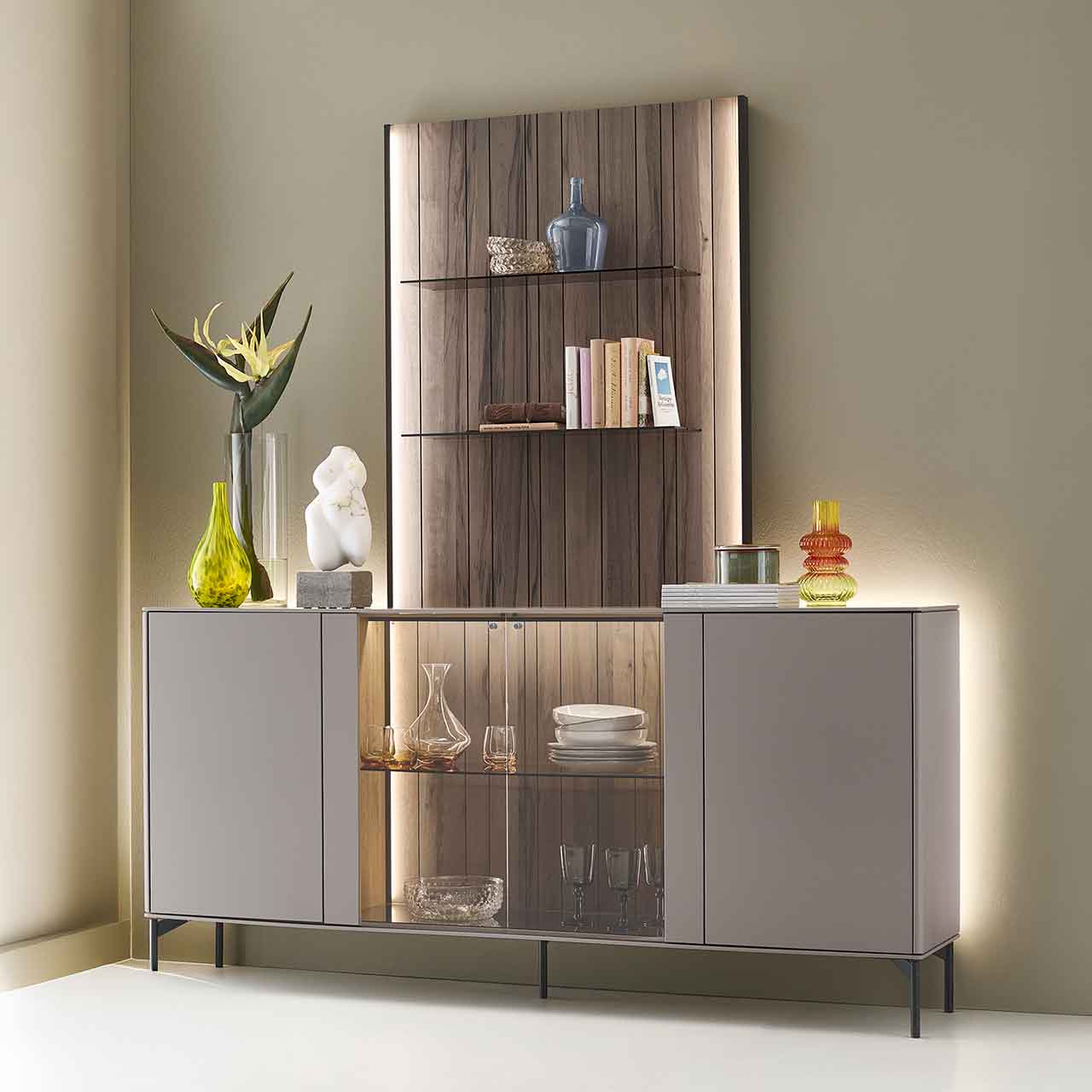 Hartmann AUDRA - Sideboard-Paneel Kombination 5610-V32 - Lack Sand und Altholzbuche Uyaa Modernes Massivholz-Sideboard auf Metallfüßen mit zwei Türen rechts und links lackiert in Sandfarben, mittig mit Glasausschnitt und Glasboden vor beleuchteter Holzrückwand in edler, dunkelbrauner Altholzbuche Uyaa, darüber Holzpaneel mit Glasböden.