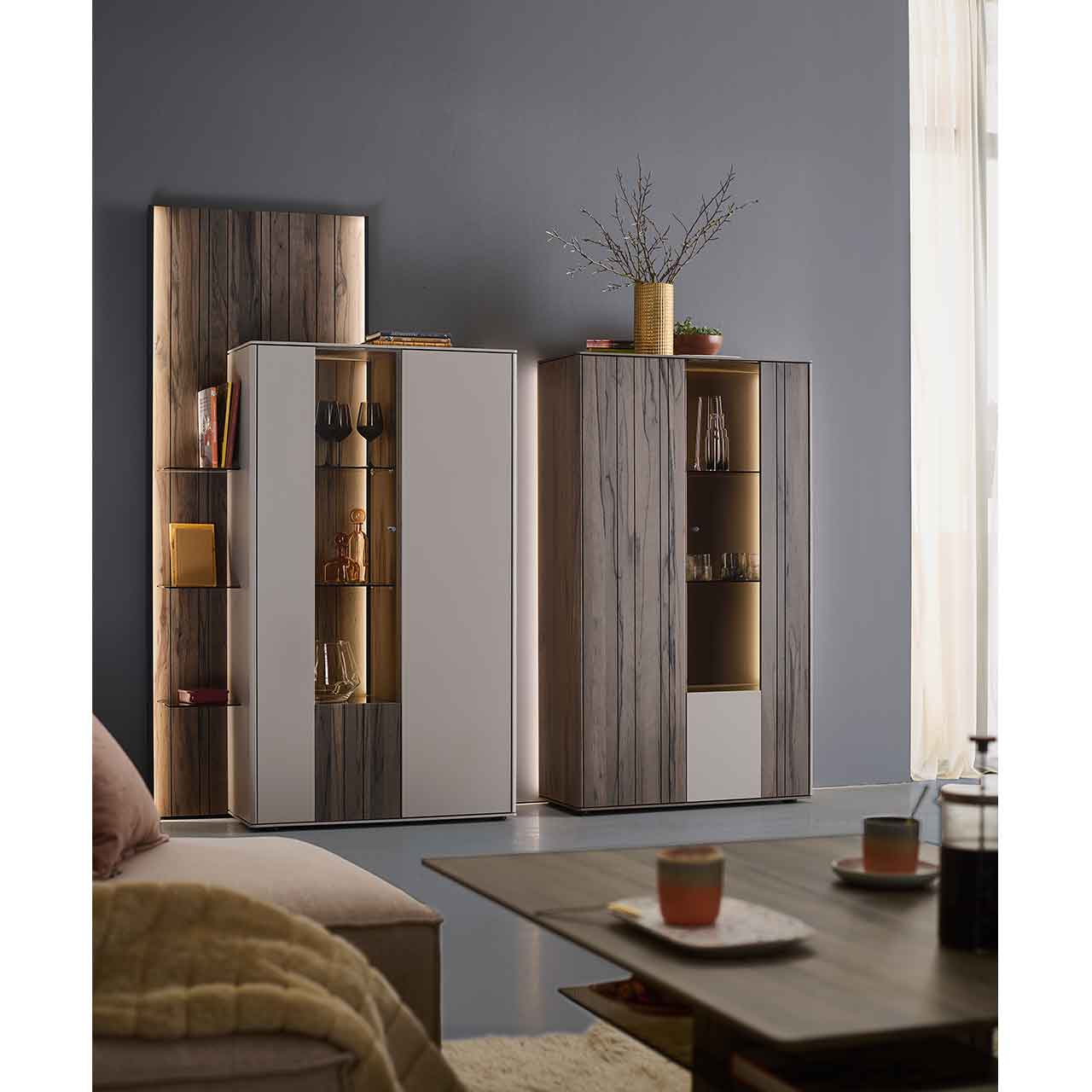 Wohnzimmer mit zwei modernen Massivholz-Highboards in sandfarbenen Lack und dunkelbrauner Altholzbuche Uyaa, jeweils mit beleuchtetem Glasausschnitt und Glasböden, ergänzt durch ein seitliches, beleuchtetes Holzwandpaneel.