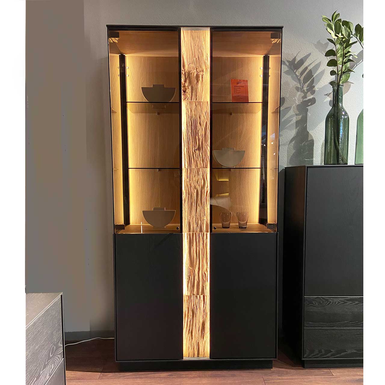 Moderne Standvitrine aus anthrazitfarbener Kernesche mit Holz,- Glastüren, beleuchteter Rückwand mit Glasböden und mittigem beleuchtetem Spaltholz-Akzent.