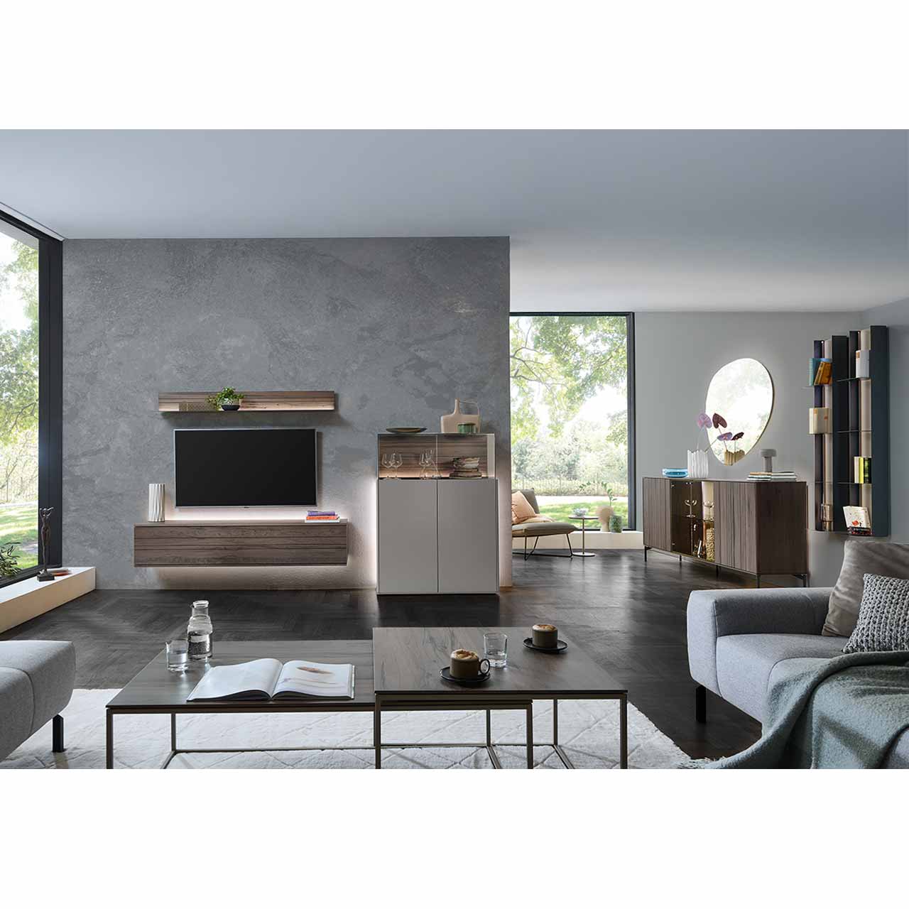 Modernes Wohnzimmer mit dreiteiliger Wohnwand bestehend aus Hängelowboard und Wandregal in dunkelbraunem Holz mit Maserung, daneben Highboard in Lack Sand mit aufgesetztem Glaskasten. Rechts an der Wand das passende Sideboard und zwei Metallregale.