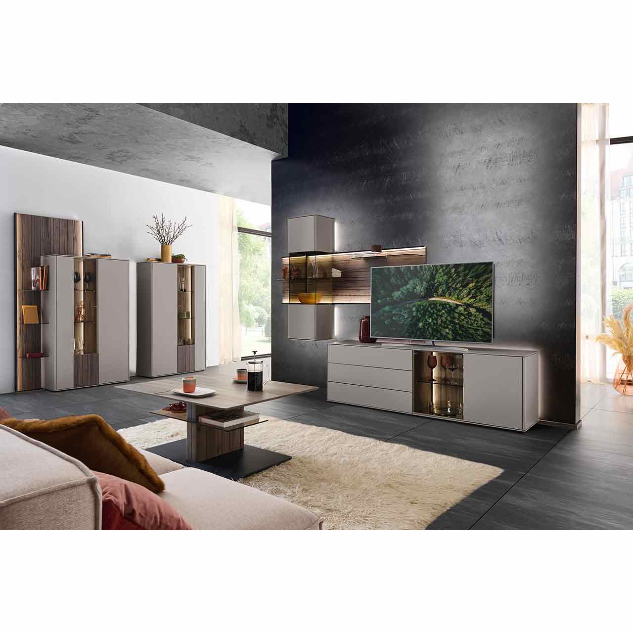 Modernes Wohnzimmer mit Wohnwand bestehend aus einem Lowboard mit Glasausschnitt und Hängevitrine in Lack Sand und beleuchtetem Wandpaneel aus Altholzbuche mit Glasböden. Davor rechteckiger Couchtisch, links zwei passende Highboards.