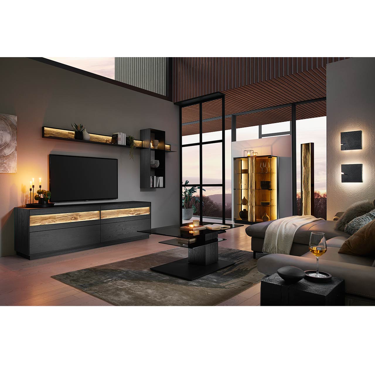 Wohnzimmer mit moderner Wohnwand in Kernesche anthrazit: Lowboard, Wandpaneel und Wandregal. Rechts das passende Highboard mit Glasfront. Im Vordergrund ein Couchtisch mit rechteckiger Glasplatte auf massiver, dunkler Holzsäule.