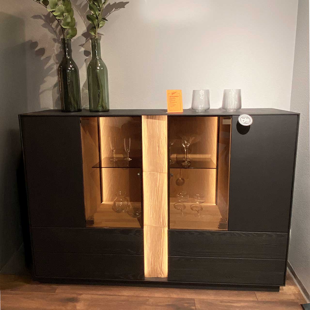 Modernes Highboard aus anthrazitfarbener Kernesche mit mittigem beleuchtetem Glasausschnitt und vertikalem Spaltholz-Akzent, innen mit Glaseinlegeböden. Links und rechts geschlossene Fronten.