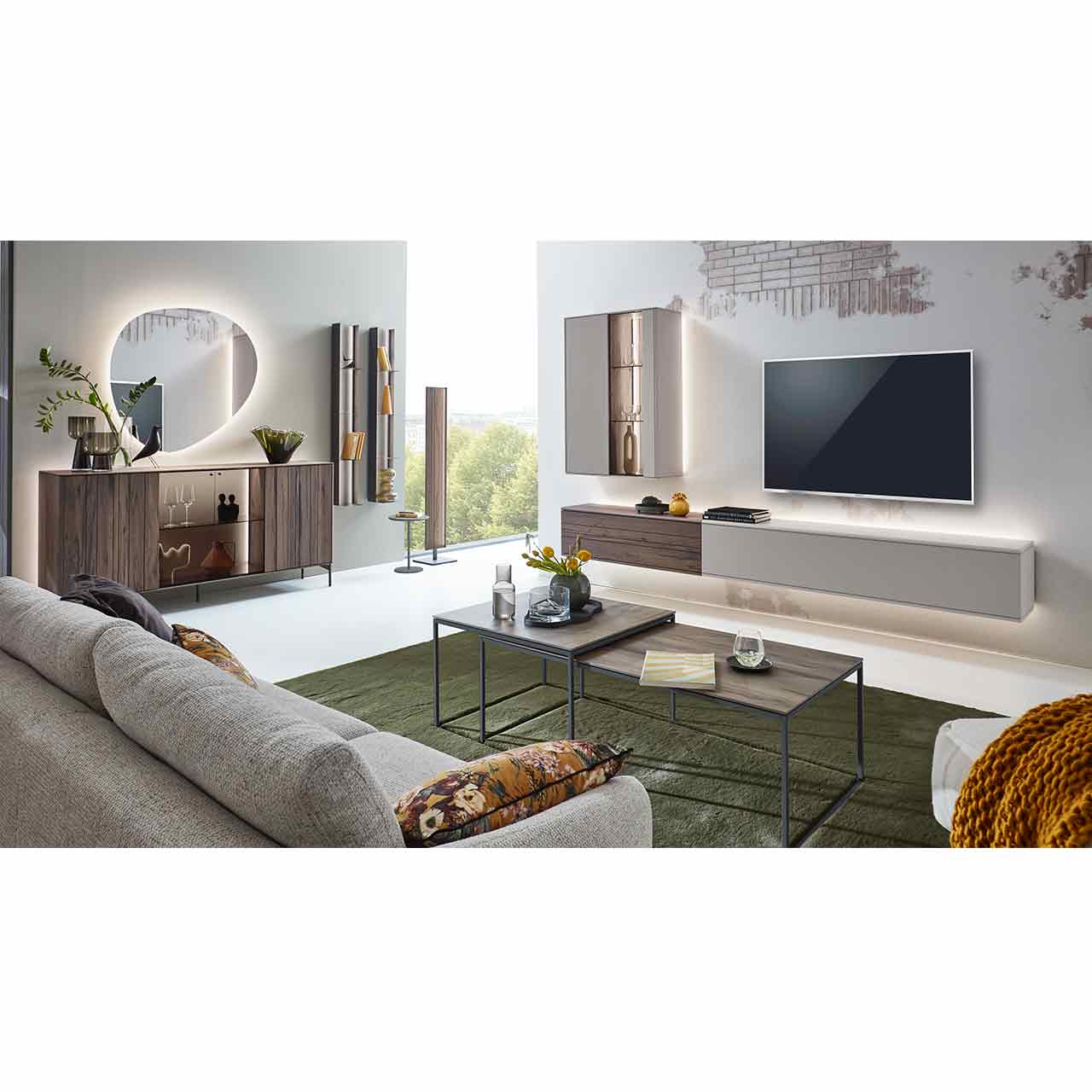 Wohnzimmer mit moderner Massivholzwohnwand bestehend aus zwei Lowboards in dunkelbraunem Holz und Lack sand und beleuchteter Hängevitrine in Lack sand. Davor zwei rechteckige Couchtische, links passendes Sideboard und zwei Wandregale.